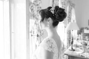 Eine Braut im Spitzenkleid steht im Profil und blickt aus einem Fenster. Ihr Haar ist elegant mit floralen Accessoires frisiert. Der Raum hinter ihr ist sanft beleuchtet und mit gemusterten Vorhängen und verschiedenen Gegenständen dekoriert.