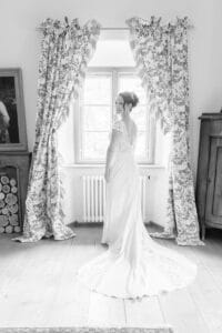 Eine Braut in einem langen, eleganten Brautkleid steht vor einem Fenster mit gemusterten Vorhängen, blickt über die Schulter zurück und lächelt. Der Raum ist hell und im klassischen Stil eingerichtet. Das Foto ist schwarz-weiß.