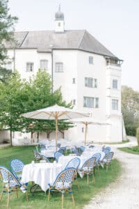 Tische im Freien mit weißen Tischdecken und blau gemusterten Stühlen stehen unter großen Sonnenschirmen auf einer Wiese vor einem großen weißen historischen Gebäude mit grünen Fensterläden und Bogenfenstern.