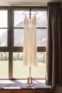 Ein langes, elegantes Brautkleid hängt an einem Fenster mit einer Berglandschaft im Hintergrund. Unter dem Kleid stehen Riemchen-Pumps auf der Fensterbank. Die Szene vermittelt eine ruhige und romantische Atmosphäre.