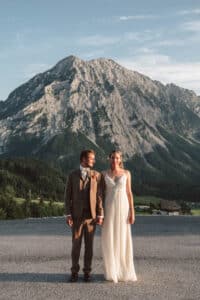 Ein Paar steht Hand in Hand vor einem großen Berg. Der Mann trägt einen braunen Anzug, die Frau ein weißes Kleid. Sie sind draußen, umgeben von klarem Himmel und viel Grün, was ein Gefühl von Ruhe und Freude vermittelt.