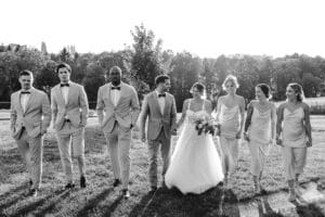 Ein Schwarz-Weiß-Foto einer Hochzeitsgesellschaft im Freien. Die Braut in einem wallenden Kleid und der Bräutigam im Anzug stehen in der Mitte, umgeben von Trauzeugen in Anzügen und Brautjungfern in Kleidern. Im Hintergrund sind Bäume und Gras zu sehen.
