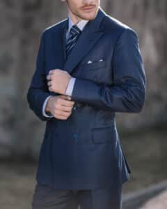 Ein Mann in einem eleganten blaukarierten Anzug und gestreifter Krawatte richtet seine Manschette, während er im Freien posiert. Er trägt ein weißes Einstecktuch und steht vor einem unscharfen Hintergrund.