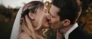 Braut und Bräutigam küssen sich im Freien. Die Braut hat langes blondes Haar und einen Schleier, der Bräutigam trägt einen dunklen Anzug. Sie sind von verschwommenem Grün umgeben, was eine romantische Atmosphäre schafft.