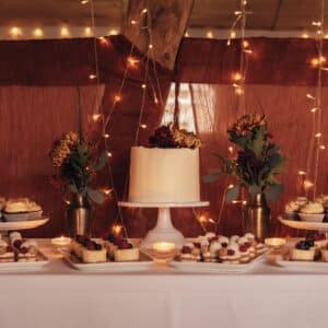 Ein Desserttisch mit einer weiß glasierten Torte auf einem Ständer, umgeben von Tellern mit Gebäck und Cupcakes. Zwei Blumenarrangements mit roten und gelben Blüten schmücken den Tisch. Im Hintergrund hängen warme Lichterketten und schaffen eine gemütliche Atmosphäre.