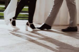 Eine Nahaufnahme von zwei nebeneinander gehenden Personen; die eine trägt weiße High Heels und ein weißes Kleid, die andere schwarze Schuhe und hellgraue Hosen. Ihre Schatten sind auf dem Boden sichtbar.