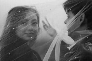 Ein Schwarzweißfoto von zwei Personen, die von einem transparenten Schleier bedeckt sind, der mit kleinen Perlen verziert ist. Eine Person blickt in die Kamera und lächelt, während die andere ihnen gegenübersteht und teilweise sichtbar ist.