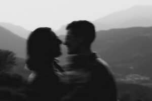 Ein unscharfes Schwarzweißfoto zeigt zwei Personen, die dicht beieinander stehen und sich gegenüberstehen. Im Hintergrund sind Berge und ein dunstiger Himmel zu sehen. Ihre Gesichter und Details sind unscharf.
