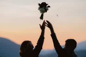 Zwei Menschen strecken gemeinsam die Hände nach einem Blumenstrauß in der Luft aus, dessen Silhouette sich vor einem sanften Sonnenuntergangshimmel mit Bergen im Hintergrund abzeichnet.