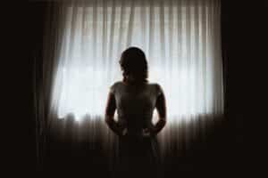 Eine Person in einem weißen Kleid steht vor einem großen Fenster mit transparenten Vorhängen. Ihre Silhouette wird durch das helle, natürliche Licht, das von hinten hereinströmt, hervorgehoben.