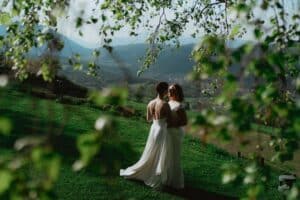 Zwei Frauen in weißen Kleidern umarmen sich auf einem grasbewachsenen Hügel, umgeben von grünen Blättern, mit Bergen und einem dunstigen Himmel im Hintergrund. Sonnenlicht fällt durch das Laub und schafft eine ruhige und romantische Atmosphäre.