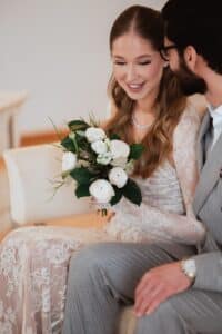 Eine Braut im Spitzenkleid lächelt und hält einen Strauß weißer Blumen in der Hand. Neben ihr sitzt ein Bräutigam im grauen Anzug. Die beiden sitzen eng beieinander auf einem hellen Sofa.
