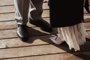 Nahaufnahme von zwei Personen, die auf einem Holzdeck stehen; eine trägt graue Hosen und schwarze Abendschuhe, die andere ein weißes Spitzenkleid und weiße spitze Pumps.