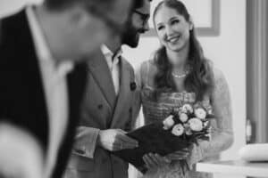 Ein lächelndes Paar steht zusammen. Die Frau hält einen Blumenstrauß, der Mann einen Ordner. Eine Person im Vordergrund ist verschwommen. Die Szene vermittelt eine warme, fröhliche Atmosphäre, vermutlich bei einer Feier oder Zeremonie.