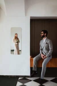 Ein Mann im grauen Anzug sitzt auf einer Bank und blickt in einen Wandspiegel, in dem sich eine Frau in einem Spitzenbrautkleid mit einem Blumenstrauß spiegelt. Der Raum hat weiße Wände und einen schwarz-weiß karierten Boden.