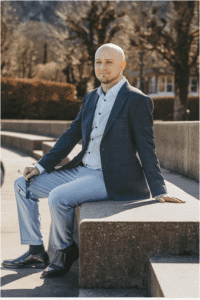 Ein kahlköpfiger Mann in blauem Blazer und heller Hose sitzt im Freien auf einer Betonbank. Er hält eine Brille in der Hand und blickt mit einem leichten Lächeln in die Kamera. Im Hintergrund sind Bäume und ein Gebäude zu sehen.