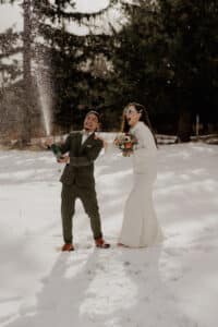 Ein fröhliches Paar in Hochzeitskleidung steht im Schnee. Der Bräutigam öffnet eine Champagnerflasche und verspritzt sie, während die Braut mit einem Brautstrauß und Sonnenbrille neben ihm lacht. Im Hintergrund sind hohe immergrüne Bäume zu sehen.