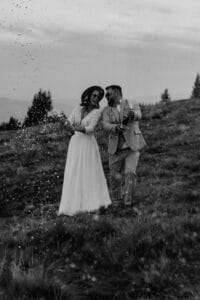 Ein fröhliches Paar in Hochzeitskleidung steht draußen auf einem grasbewachsenen Hügel, lässt Champagner knallen und lacht gemeinsam, während die Gischt um sie herum spritzt. Das Bild ist in Schwarzweiß.
