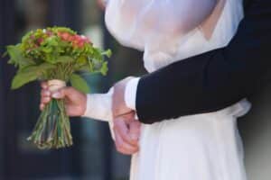 Eine Nahaufnahme von Braut und Bräutigam, die sich umarmen. Die Braut im weißen Kleid hält einen Strauß grüner und orangefarbener Blumen, während der Bräutigam im dunklen Anzug sanft ihre Taille hält.