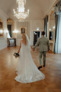 Eine Braut in einem weißen Kleid und Schleier hält einen Blumenstrauß und geht Hand in Hand mit einem Bräutigam in einem hellen Anzug durch einen eleganten, von einem Kronleuchter erleuchteten Ballsaal mit kunstvollem Dekor.