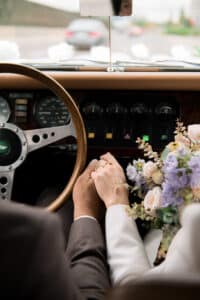 Ein Paar hält Händchen in einem Oldtimer, wobei eine Person einen pastellfarbenen Blumenstrauß hält. Durch die Windschutzscheibe sind Armaturenbrett, Lenkrad und die verschwommene Straße zu sehen.