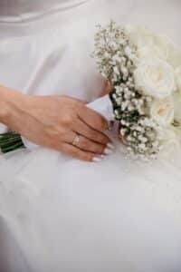 Eine Nahaufnahme einer Hand mit einem goldenen Ring, die einen Strauß weißer Rosen und Schleierkraut hält und auf einem weißen Brautkleid ruht, das mit kleinen Perlen verziert ist.