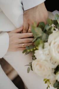 Nahaufnahme zweier Hände mit goldenen Eheringen, die sich sanft berühren, neben einem Strauß weißer Rosen und grüner Blätter, die eine Hochzeit oder romantische Verbindung symbolisieren.