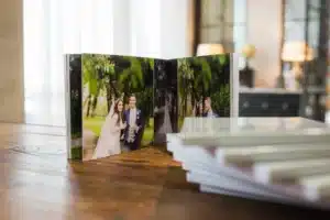 Auf einem Holztisch liegt ein offenes Fotoalbum mit Hochzeitsfotos eines lächelnden Brautpaares im Freien. Daneben stapeln sich mehrere geschlossene Fotoalben. Im Hintergrund ist ein sanft beleuchteter Raum zu sehen.