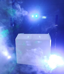 Ein DJ-Pult mit blauen und grünen Lichtern, die durch den Nebel scheinen und eine lebendige und energiegeladene Atmosphäre schaffen. Zwei Lautsprecher stehen auf beiden Seiten des Pults, Lichtstrahlen und Rauch erfüllen die Szenerie.