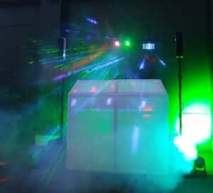 Bunte Laserlichter und Nebel umgeben eine DJ-Kabine mit Lautsprechern auf beiden Seiten und schaffen eine lebhafte Partyatmosphäre in einem dunklen Innenbereich.