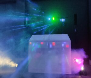 Bunte Bühnenlichter und Laserstrahlen durchdringen dichten Nebel und beleuchten ein mit weißem Tuch bedecktes DJ-Pult. Der dunkle Hintergrund verstärkt die lebendigen Effekte und die energiegeladene Atmosphäre.