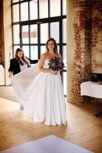 Eine Braut in einem weißen Kleid hält einen Strauß lila Blumen, während eine Frau hinter ihr ihr Kleid zurechtrückt. Sie stehen in einem rustikalen Raum mit Backsteinwänden und großen Fenstern.