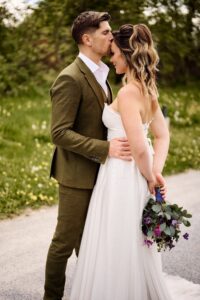 Ein Bräutigam im grünen Anzug küsst die Stirn seiner Braut, die ein weißes Kleid trägt und einen Strauß lila Blumen in der Hand hält. Sie stehen dicht beieinander auf einem Weg im Freien, im Hintergrund ist Grün.