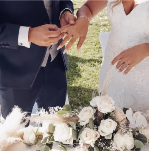 Der Bräutigam steckt der Braut während einer Hochzeitszeremonie im Freien einen Ring an den Finger. Die Braut trägt ein weißes Spitzenkleid, und auf dem Tisch vor ihnen liegt ein Strauß weißer Rosen und Grünzeug.