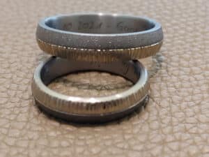 Zwei metallische Ringe mit strukturierten Kanten sind auf einer beigen, lederähnlichen Oberfläche übereinander angeordnet. In den inneren Teil der Ringe scheint ein Text eingraviert zu sein.