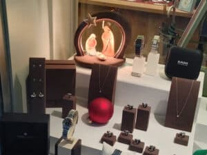 Schmuckauslage mit Halsketten, Ringen und Uhren. Im Vordergrund steht ein rotes Ornament. Im Hintergrund befindet sich eine beleuchtete Krippendekoration mit den stilisierten Figuren von Maria, Josef und dem Jesuskind.