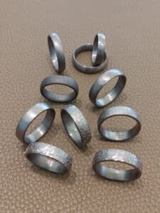 Zehn Metallringe mit unterschiedlichen Strukturen und Oberflächen sind auf einer braunen, strukturierten Oberfläche angeordnet. Einige Ringe sind glatt, andere haben ein gehämmertes Muster.