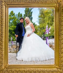 Eine Braut in einem weißen Kleid und ein Bräutigam in einem dunklen Anzug mit Stock stehen zusammen im Freien, eingerahmt von einem verzierten goldenen Rahmen, mit Bäumen und lila Blumen im Hintergrund.