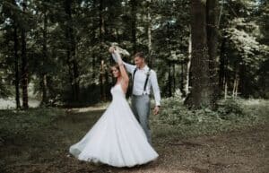 Eine Braut in einem weißen Kleid wirbelt fröhlich herum, während der Bräutigam, der ein weißes Hemd und Hosenträger trägt, ihre Hand in einer bewaldeten Umgebung mit Bäumen und sanftem Sonnenlicht im Hintergrund hält.