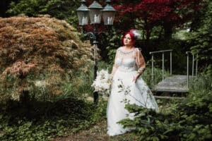 Eine Braut mit rotem Haar und weißem Kleid steht im Freien in einem üppigen, grünen Garten mit einem Strauß weißer Blumen in der Hand, in der Nähe eines alten Laternenpfahls und einer kleinen Holzbrücke.
