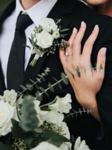 Nahaufnahme eines Paares in förmlicher Kleidung, wobei eine Person einen schwarzen Anzug, ein weißes Hemd und eine schwarze Krawatte trägt, die mit einer weißen Rosenboutonniere geschmückt ist. Die manikürte Hand der anderen Person, die einen Verlobungsring trägt, ruht auf dem Anzug.