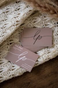 Ein Stapel brauner Visitenkarten mit eleganter weißer und schwarzer Schrift liegt auf einem cremefarbenen Strickstoff, der auf einer Holzfläche platziert ist. Auf einigen Karten sind Kontaktdaten angegeben, auf anderen nur ein stilisiertes "lp"-Logo.