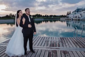 Eine Braut in einem weißen Kleid und ein Bräutigam in einem schwarzen Anzug stehen zusammen auf einem hölzernen Steg an einem ruhigen See, mit einem großen Gebäude, Bäumen und einem bewölkten Himmel im Hintergrund.