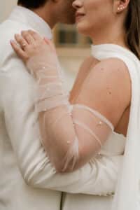 Eine Braut in einem weißen Kleid mit durchsichtigen, perlenverzierten Handschuhen umarmt einen Bräutigam in einem weißen Anzug. Die Hand der Braut ruht auf der Schulter des Bräutigams und zeigt ihren Verlobungsring, während sie einen intimen, engen Moment miteinander verbringen.