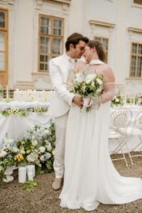 Ein weiß gekleidetes Brautpaar umarmt sich und berührt lächelnd die Stirn vor einem eleganten Gebäude und einem mit weißen und pastellfarbenen Blumen dekorierten Hochzeitstisch im Freien.