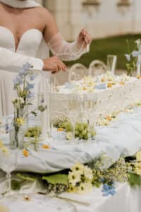 Eine Person in einem weißen, schulterfreien Kleid schneidet eine große rechteckige, mit Blumen verzierte Torte an. Der Tisch ist mit weißen und gelben Blumen, blauem Tuch, Kerzen, Weintrauben und Sektgläsern geschmückt.