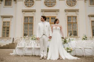 Eine Braut und ein Bräutigam in weißer Kleidung stehen händchenhaltend vor einem eleganten Gebäude mit großen Fenstern, dekorativen Stühlen und Blumenarrangements, die für eine Hochzeitszeremonie im Freien vorbereitet sind.