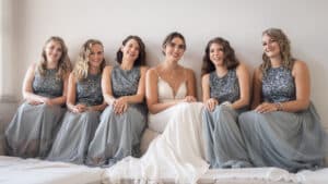 Eine Braut in einem weißen Kleid sitzt lächelnd auf einem Sofa, umgeben von fünf Brautjungfern in passenden glitzernden blau-grauen Kleidern, die alle eng beieinander sitzen und glücklich aussehen.