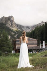 Eine Frau in einem weißen Kleid steht auf einer Wiese und blickt auf ein hölzernes Gebäude, das sich unter einem wolkenverhangenen Himmel gegen hohe, felsige Berge und Kiefern abhebt.