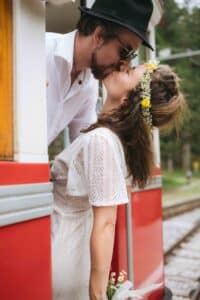 Ein Paar küsst sich, während es sich aus einer roten Straßenbahn lehnt. Die Frau, die ein weißes Spitzenkleid und eine Blumenkrone trägt, hält Blumen in der Hand. Der Mann trägt eine Sonnenbrille, ein weißes Hemd und einen schwarzen Hut. Im Hintergrund sind Bäume zu sehen.